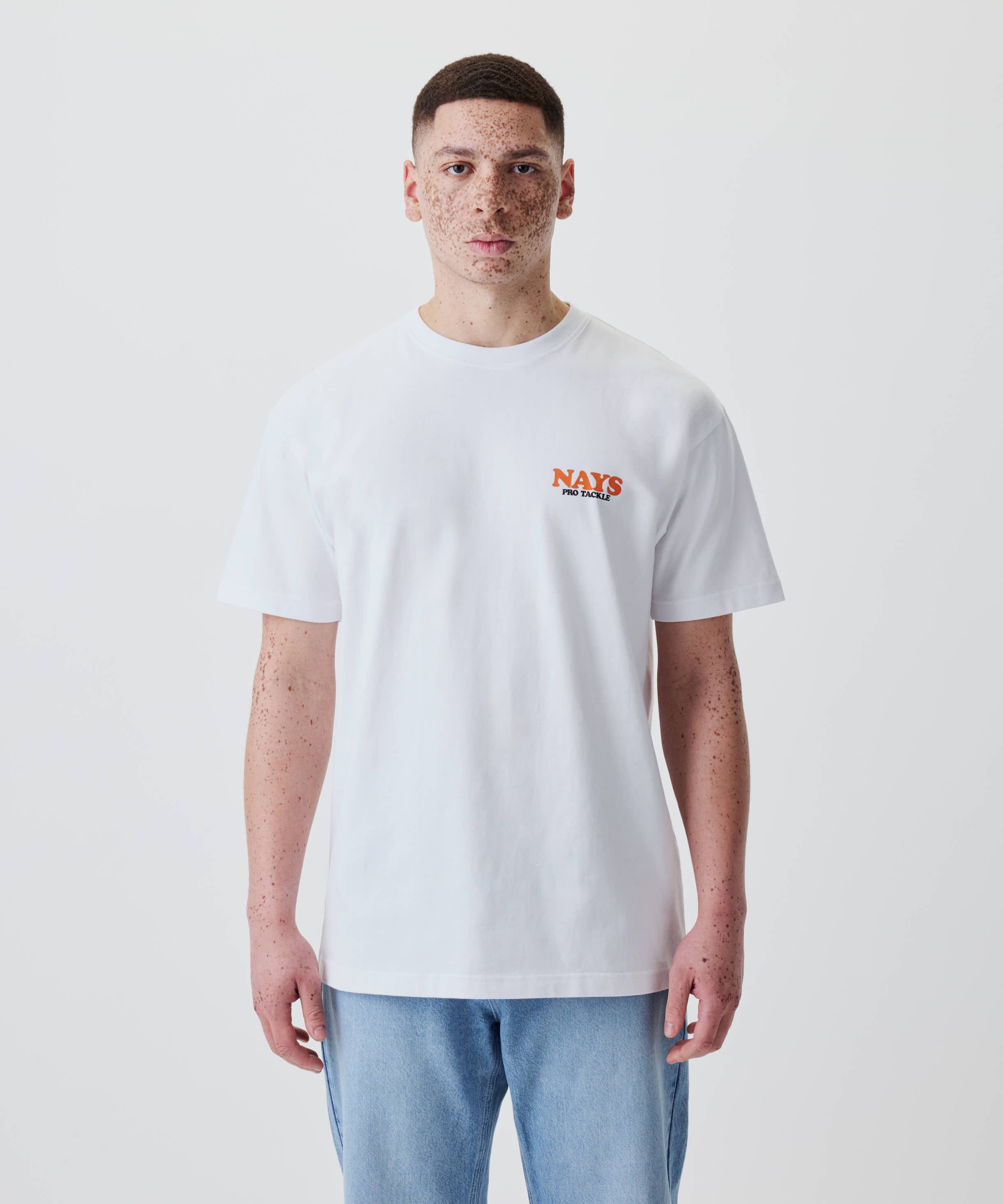 Junger Mann in einem weißen Nays College Print Tee mit kleinem Logo auf der Brust, neutraler Blick, trägt blaue Jeans, schlichtes, modernes Design.