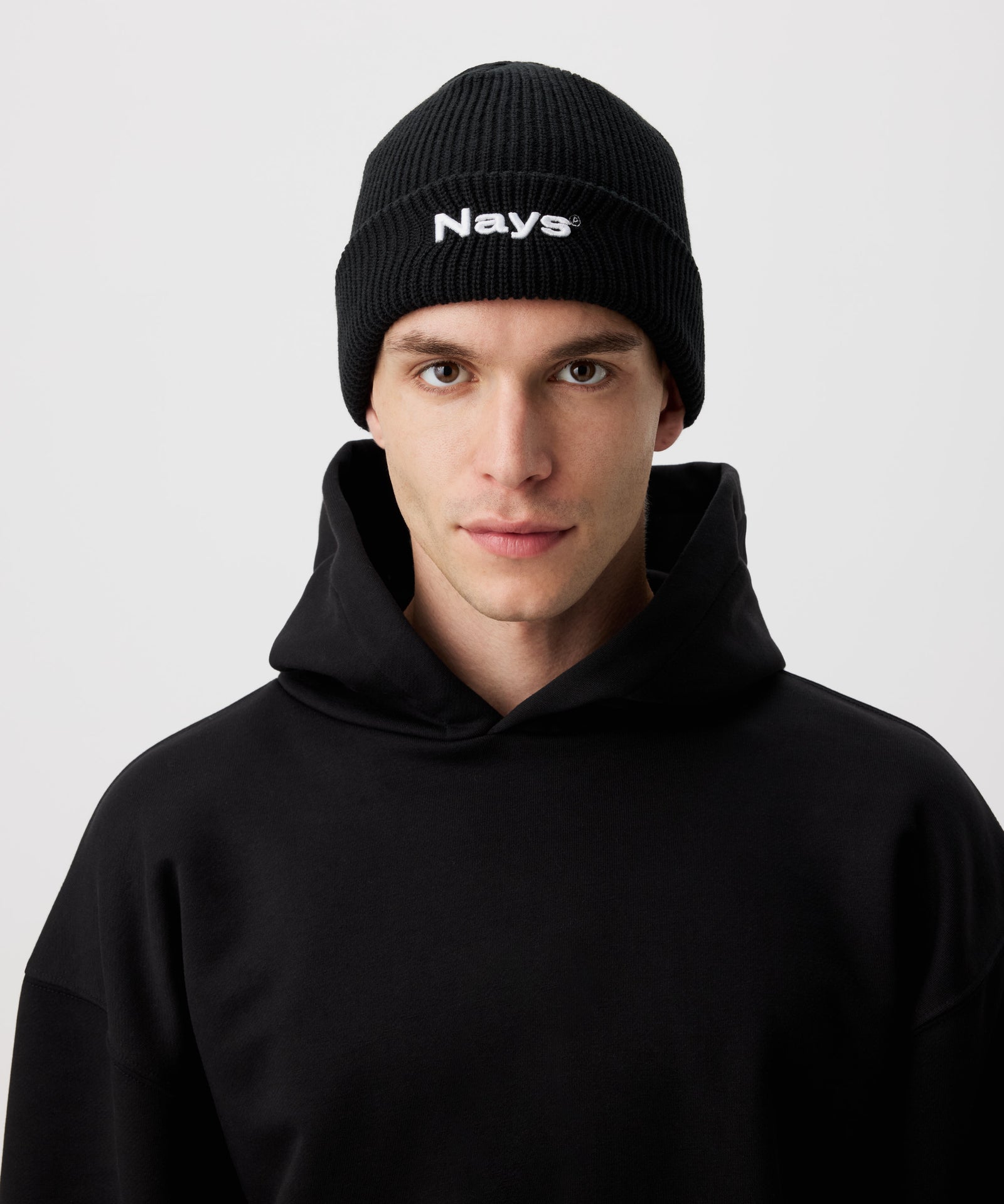 Eine schwarze Beanie Mütze, mit Nays Stickerei in weiß, getragen von einem Mann in schwarzem Hoodie.