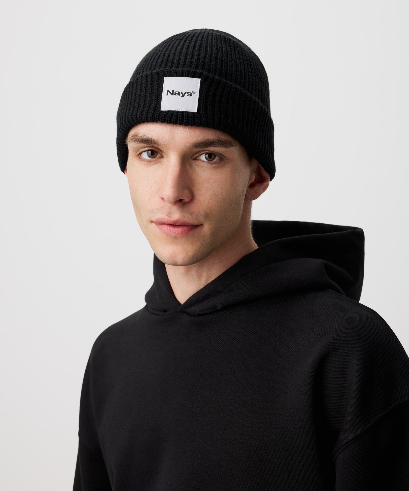 Eine schwarze Beanie Mütze mit Nays Brand Patch, getragen von einem Mann der in die Kamera schaut. 