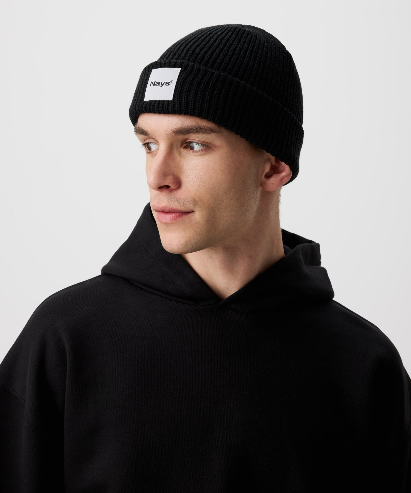 Eine schwarze Beanie Mütze mit Nays Brand Patch, getragen von einem Mann, er ist seitlich ausgerichtet.