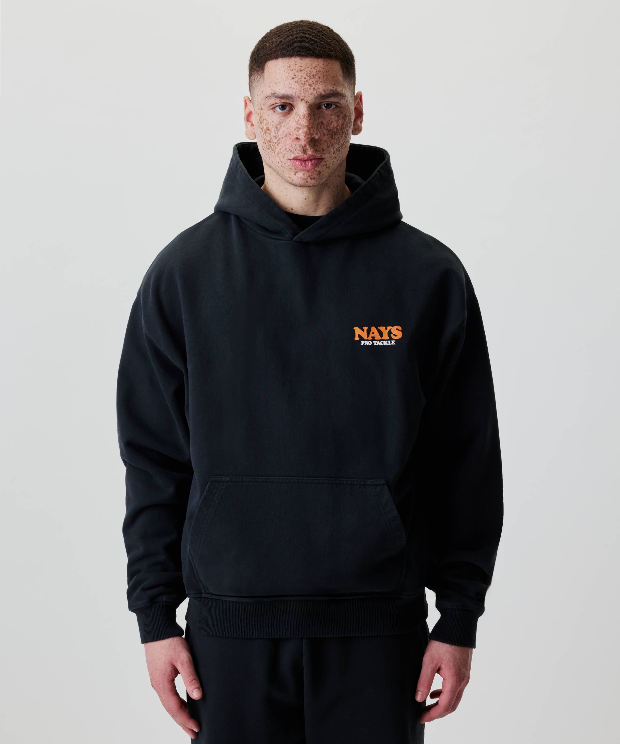 Eine Person trägt den Nays College Print Hoodie mit NAYS PRO TACKLE-Logo auf der Brust, in modernem Stil.