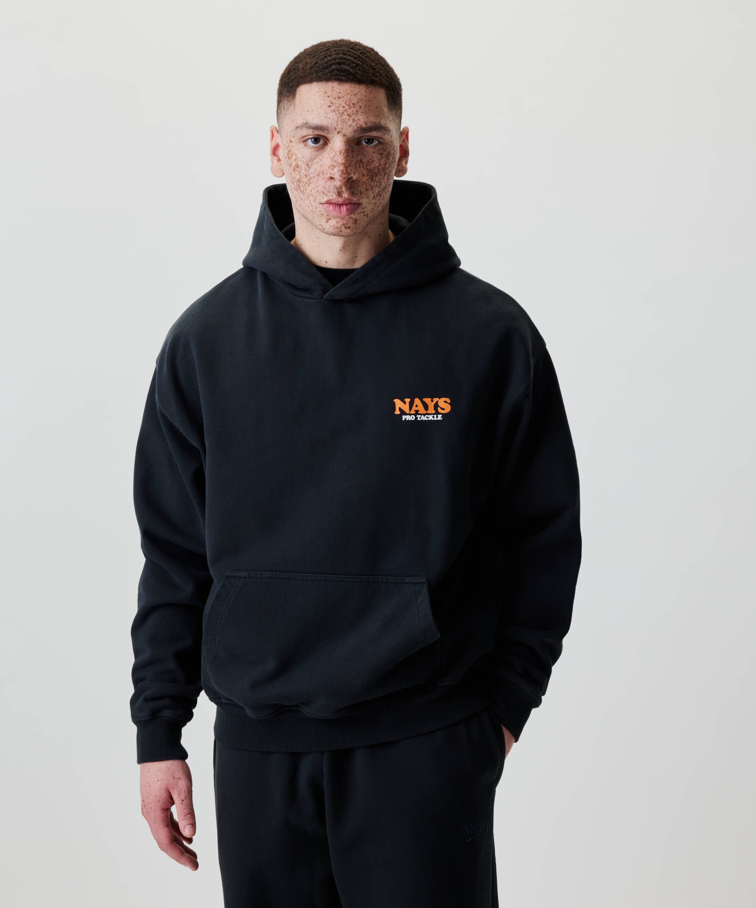 Ein junger Mann trägt das Nays College Print Hoodie in Schwarz mit orangefarbenem Logo auf der Brust, kombiniert mit passenden Sweatpants.
