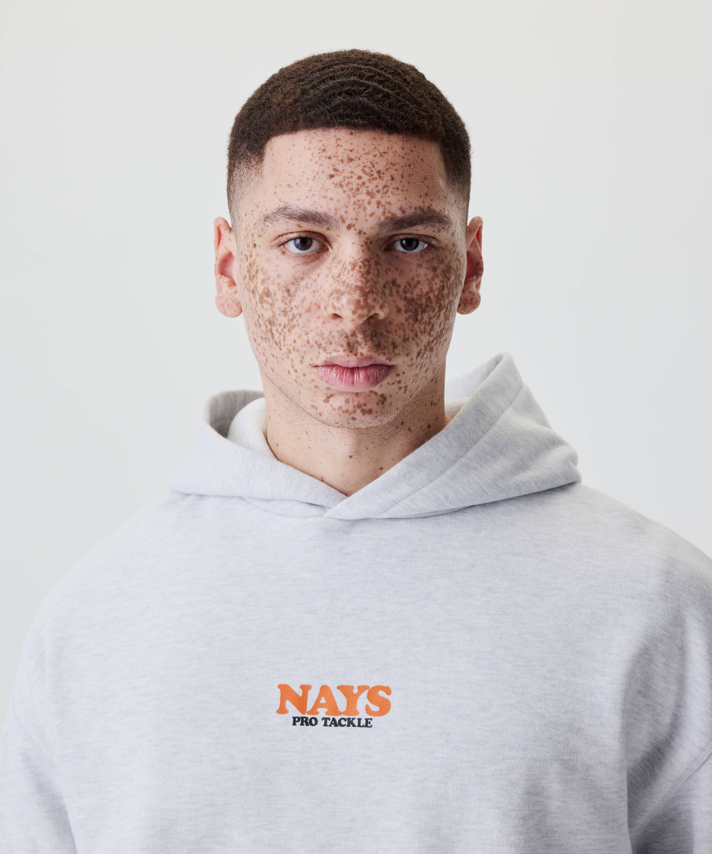Junger Mann mit Sommersprossen trägt den Nays College Print Hoodie in Grau Melange mit auffälligem NAYS PRO TACKLE-Print auf der Brust.
