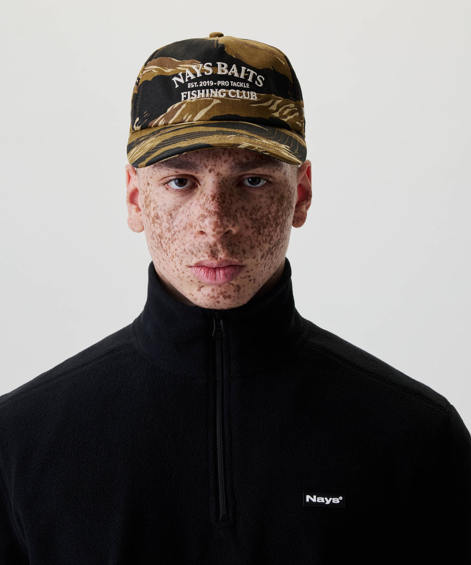 Nahaufnahme einer Person mit Nays Fishing Club Cap, die ein markantes Camo-Muster und NAYS BAITS Stickerei trägt, kombiniert mit verstellbarem Snapback-Verschluss.