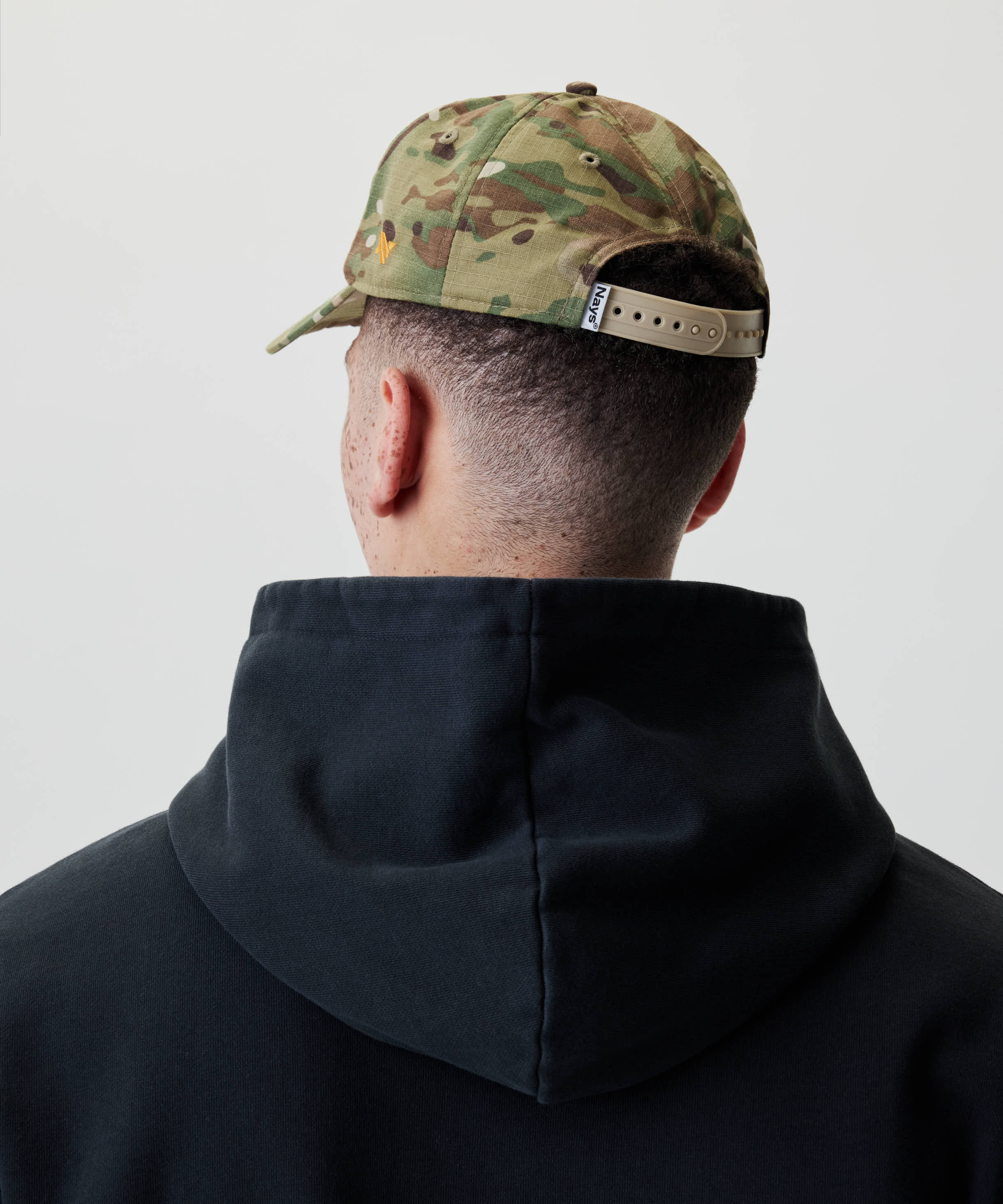 Rückansicht einer Person mit der Nays Fishing Club Cap, erkennbar am Camouflage-Muster und verstellbarem Snapback. Kombiniert mit einem dunklen Hoodie.