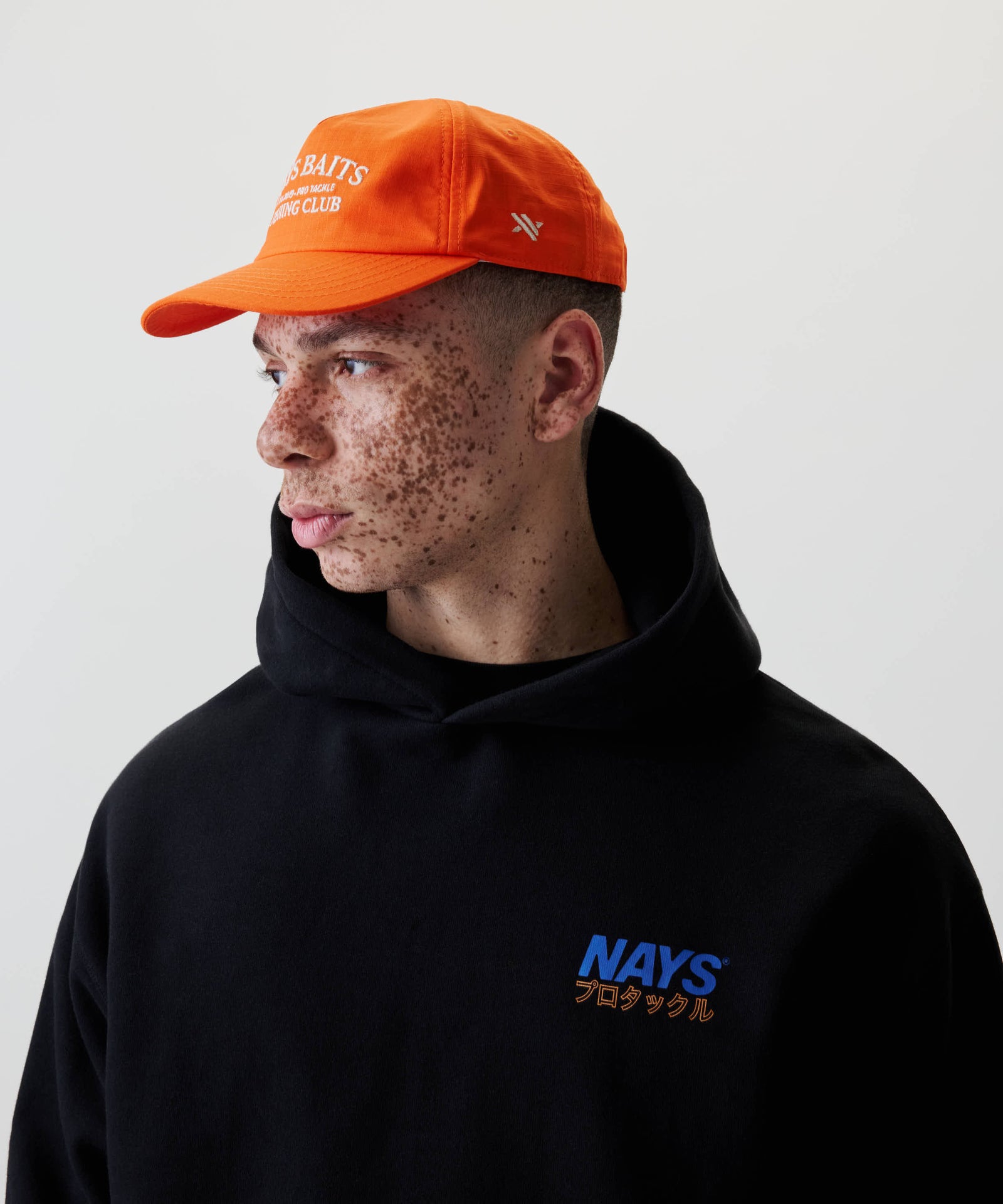 Junger Mann im Profil trägt eine leuchtend orangefarbene Baseballkappe mit weißer Aufschrift, genannt Fishing Club Cap, kombiniert mit einem schwarzen Hoodie mit NAYS-Logo.