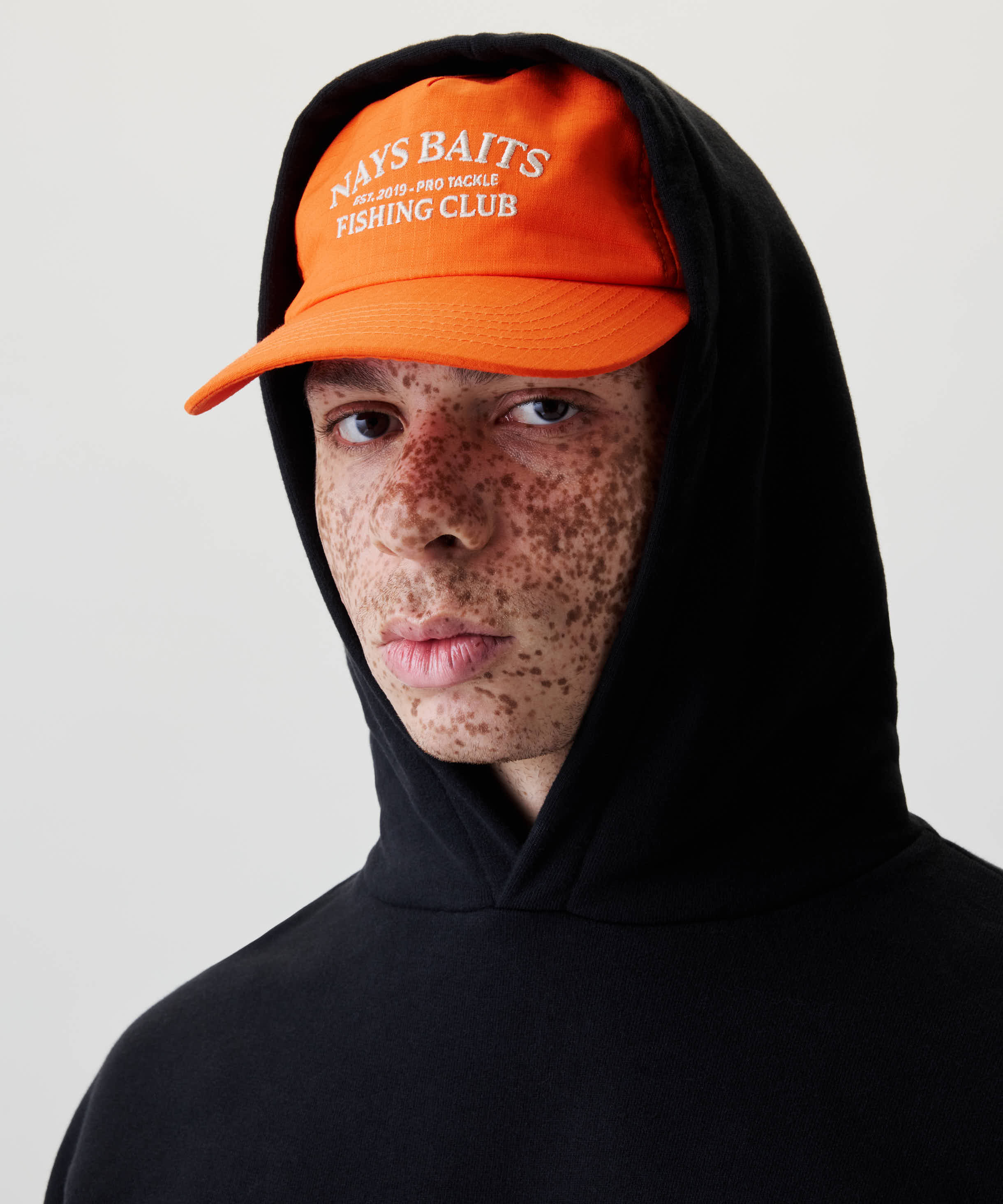 Nahaufnahme eines jungen Menschen mit einem schwarzen Kapuzenpullover und einem orangefarbenen Baseball-Cap mit der Aufschrift NAYS BAITS und FISHING CLUB.