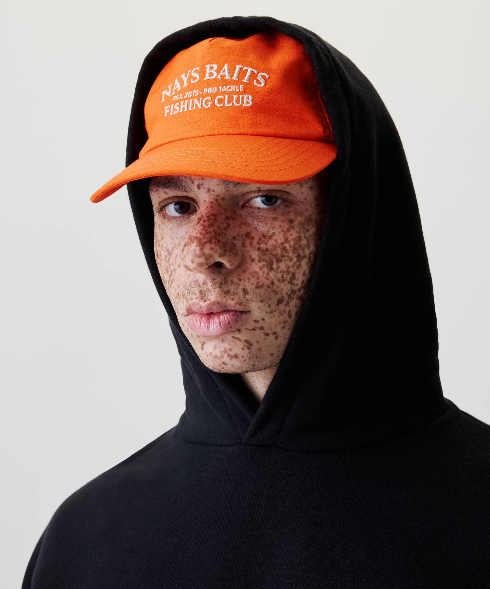 Nahaufnahme eines jungen Menschen mit einem schwarzen Kapuzenpullover und einem orangefarbenen Baseball-Cap mit der Aufschrift NAYS BAITS und FISHING CLUB.