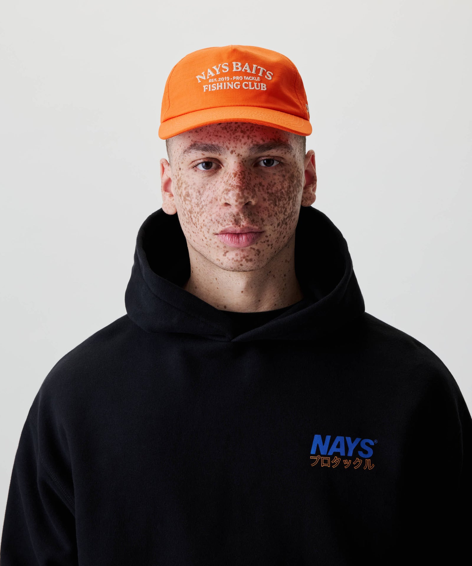 Junger Mann trägt die Fishing Club Cap von NAYS mit weißem Aufdruck, kombiniert mit einem schwarzen Hoodie, der ein kleines NAYS-Logo mit japanischen Schriftzeichen zeigt.