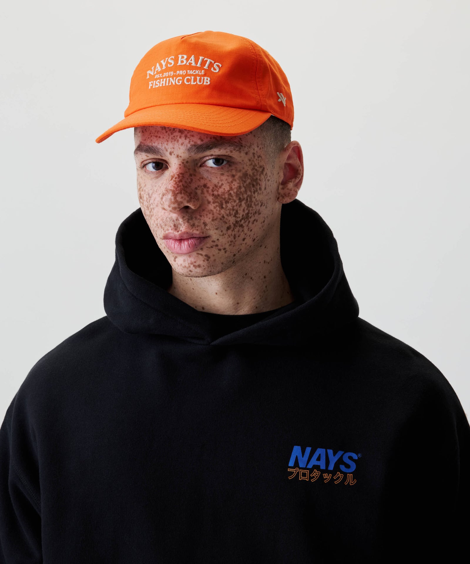 Junger Mensch trägt die Fishing Club Cap mit NAYS BAITS Stickerei und einem schwarzen Hoodie mit blauem Logo und japanischen Schriftzeichen vorne.