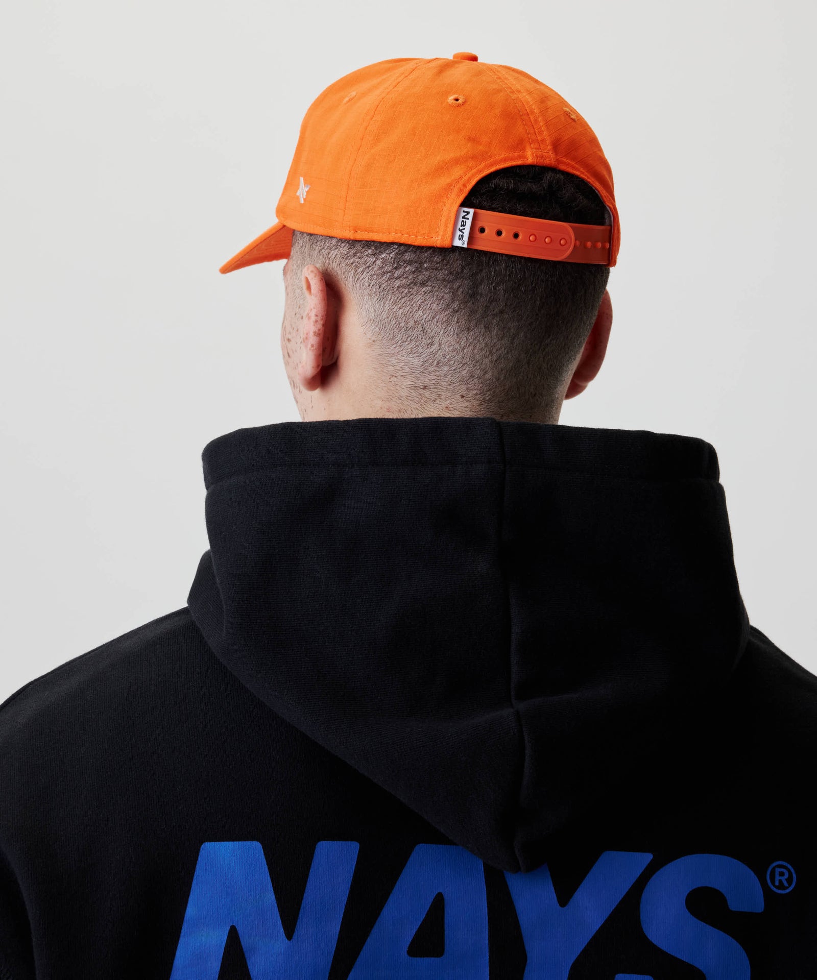 Person von hinten mit einem rückwärts getragenen Fishing Club Cap, das „Nays“ an der Verschlusslasche zeigt, und einem Hoodie mit „NAYS“-Schriftzug am unteren Rücken.