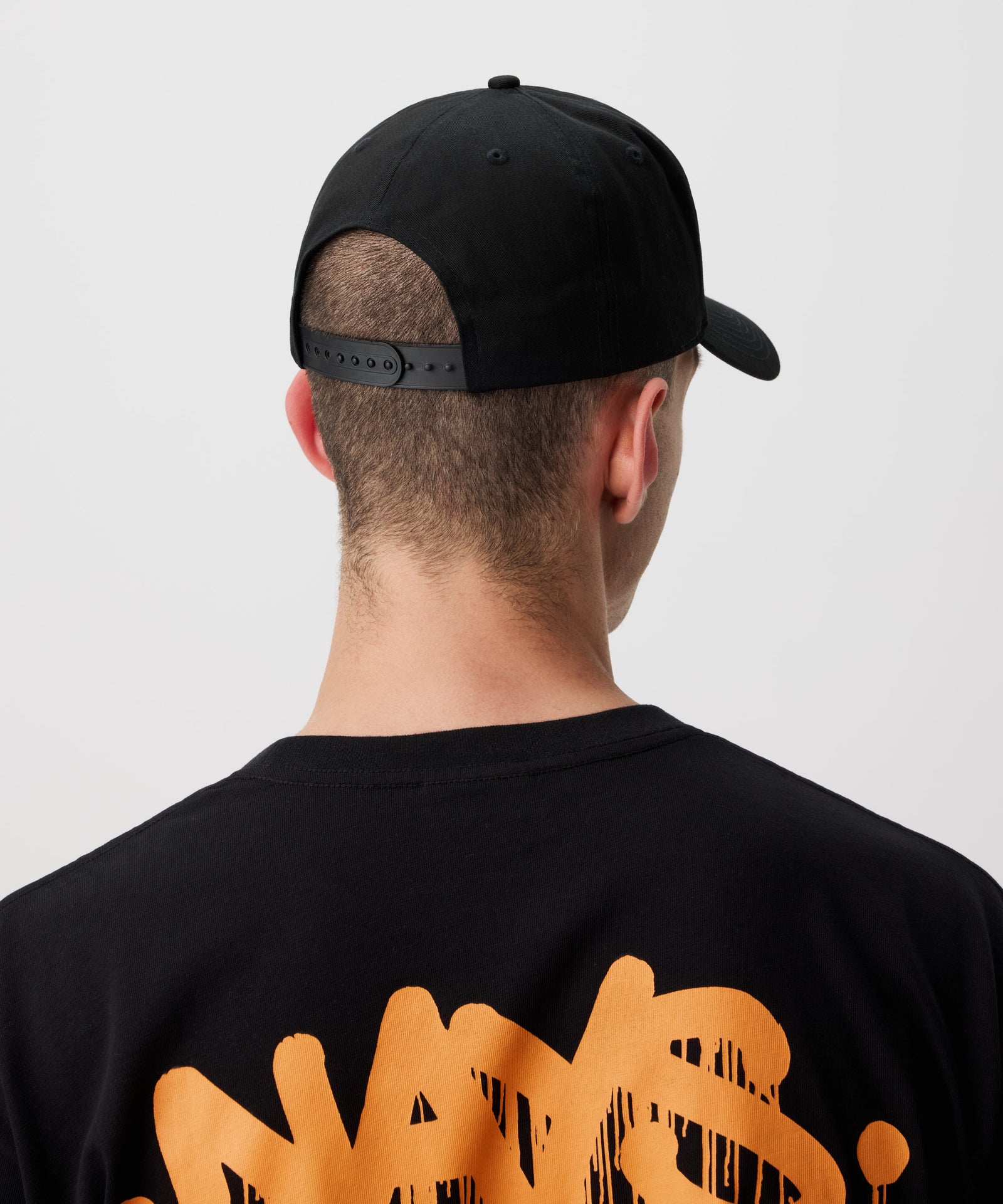 Zu sehen ist ein Mann von hinten, der eine schwarze Schirmmütze trägt.  Er trägt ein schwarzes T-Shirt mit orange-farbigem Nays Logo in Graffiti Optik.