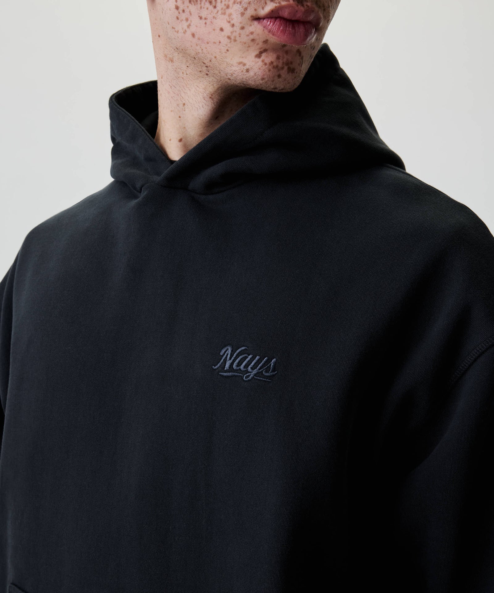Person trägt den Nays New Script Hoodie mit dezentem Logo auf der Brust. Kapuze hinten gefaltet. Baumwollmaterial für angenehmes Tragegefühl.