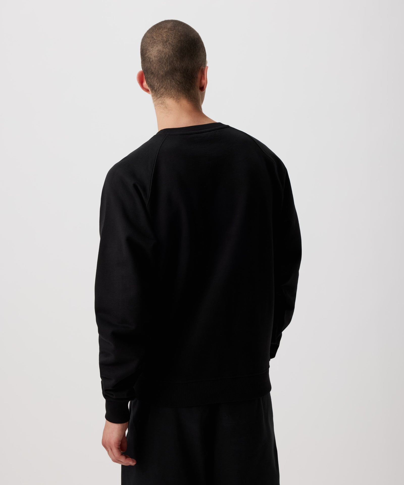 Zu sehen ist ein Mann von hinten. Er trägt einen schwarzen Pullover und eine lange, schwarze Hose.