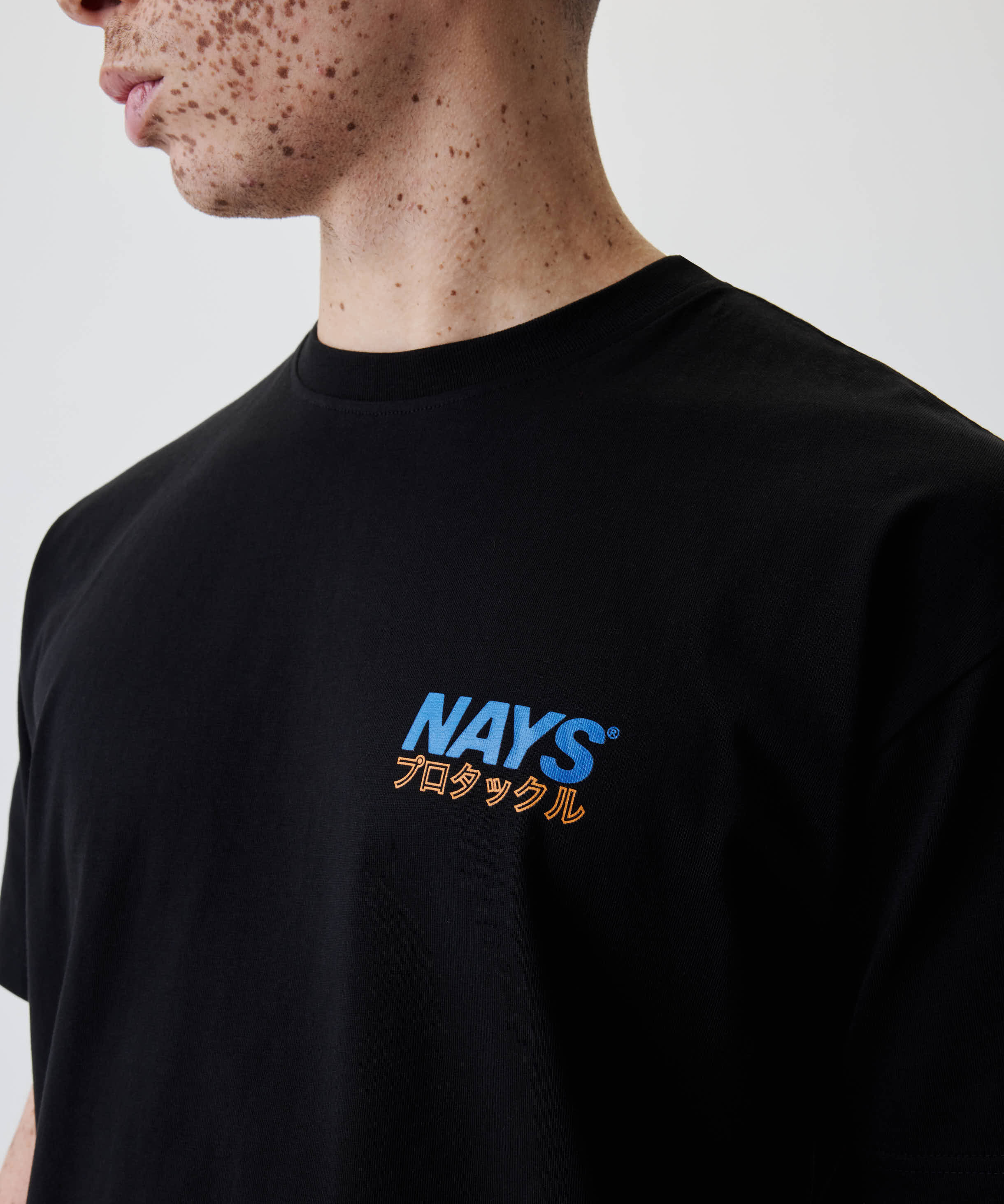 Person trägt das Nays Pro Tackle International Tee mit blauem NAYS-Logo und orangenen Katakana-Zeichen auf der Brust, aus 100% Baumwolle.