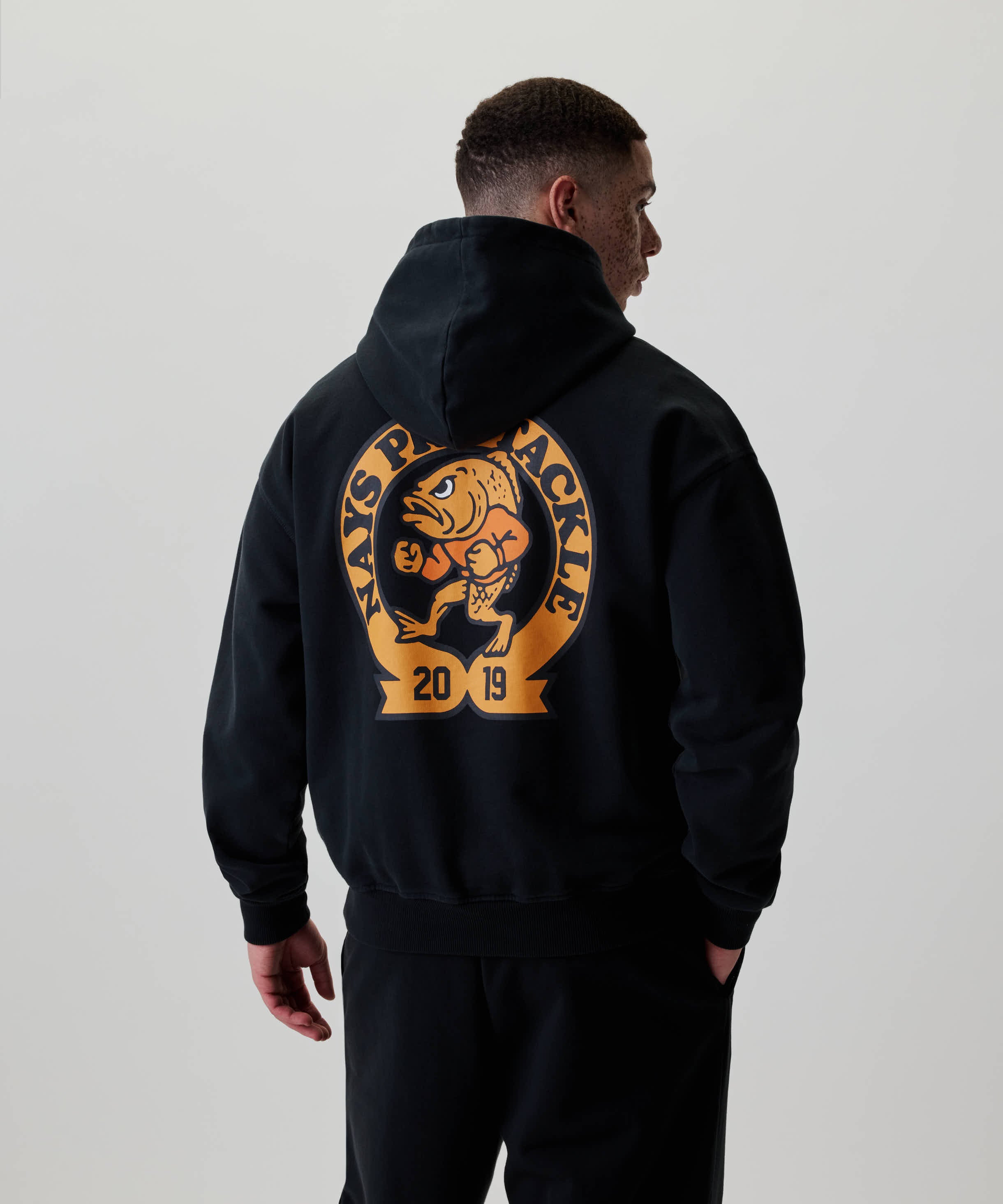 Ein junger Mann trägt den Nays College Print Hoodie mit großem Fisch-Emblem auf dem Rücken, lässig und modern für Alltag und Freizeit.