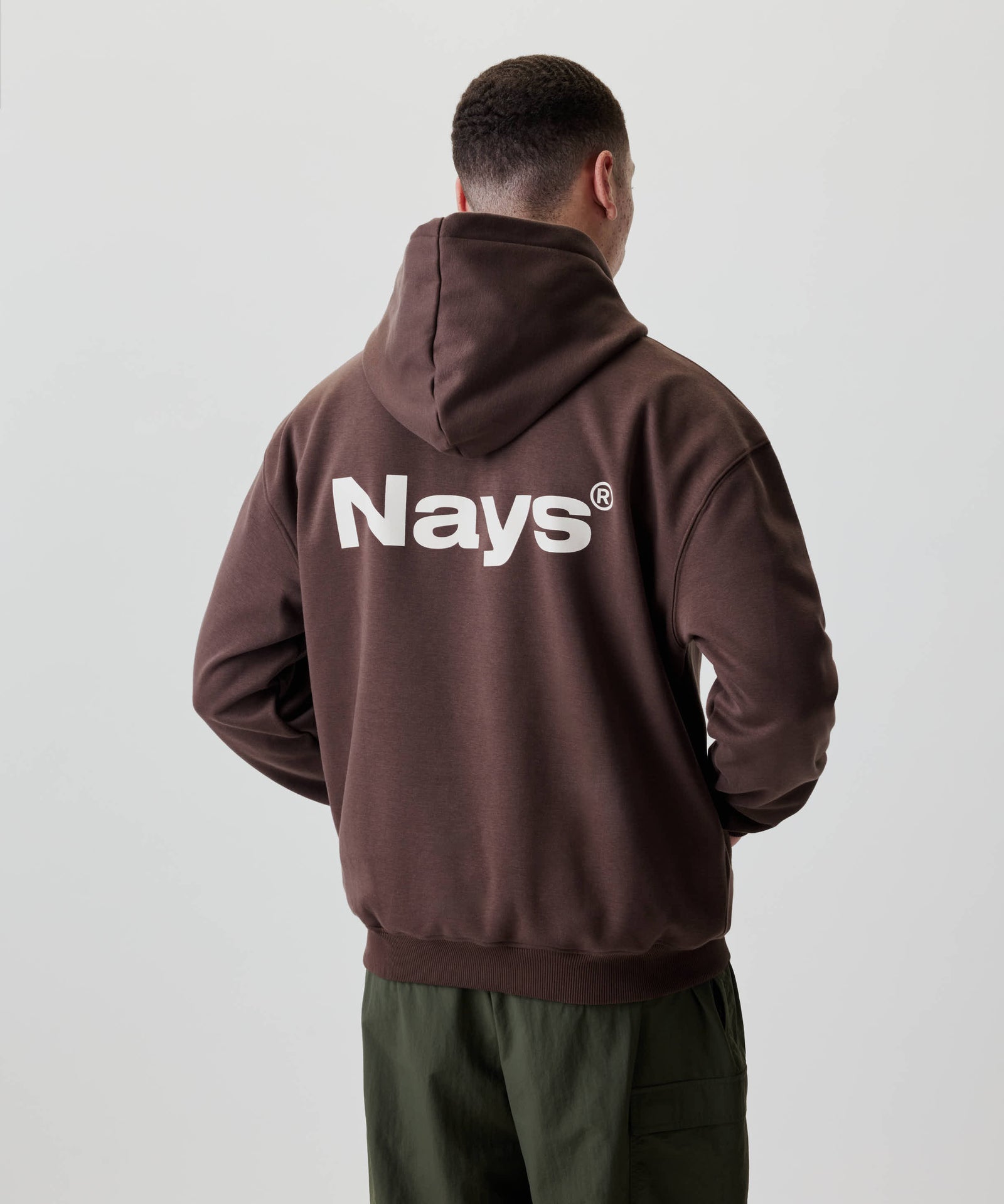 Mann von hinten mit dem Nays Quote Hoodie in Braun, auffälliger Nays®-Schriftzug in Weiß, kombiniert mit olivgrünen Cargo-Hosen, betont den lässigen Stil.