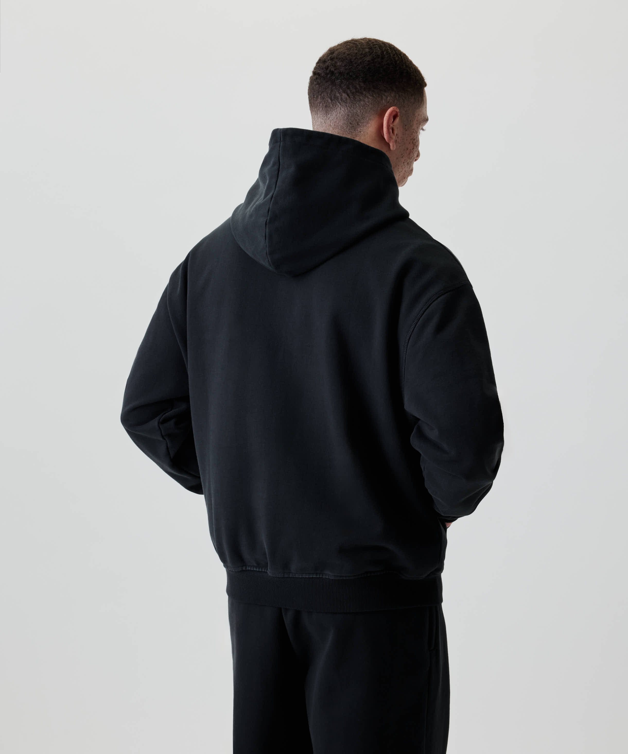 Person von hinten in einem schlichten schwarzen Nays New Script Hoodie, Hände in den Taschen, vor hellem Hintergrund. Dezente Logo-Stickerei auf der Brust.