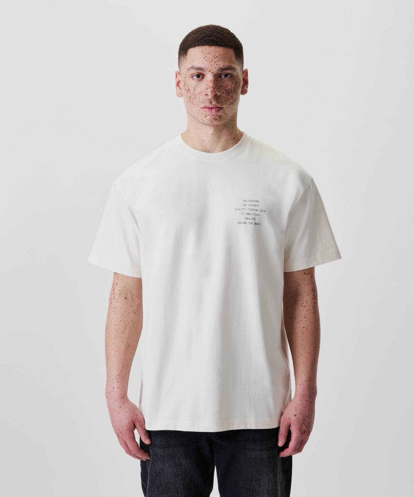 Junger Mann trägt das Nays Quote Tee in Off White mit dunkelgrünem Grafik-Print, lockerem Schnitt, kombiniert mit dunklen Jeans für einen entspannten Look.