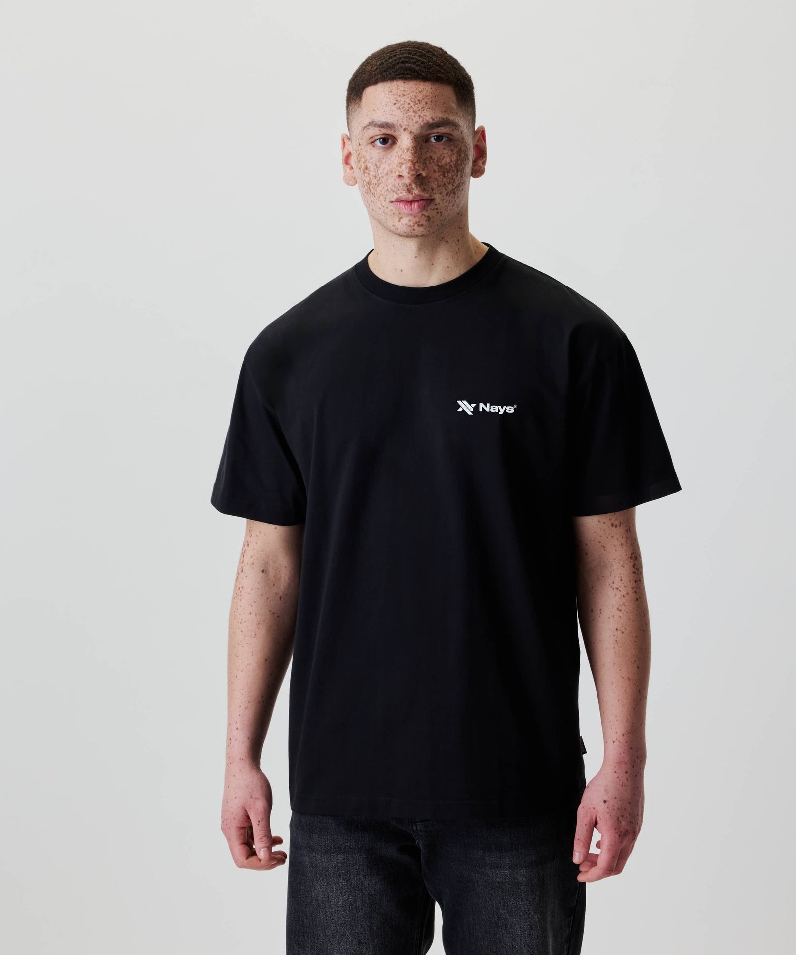 Junger Mann trägt das Nays Loose Fit Chest Print Tee in Schwarz mit weißem Logo-Print auf der Brust, kombiniert mit dunklen Jeans, vor neutralem Hintergrund.