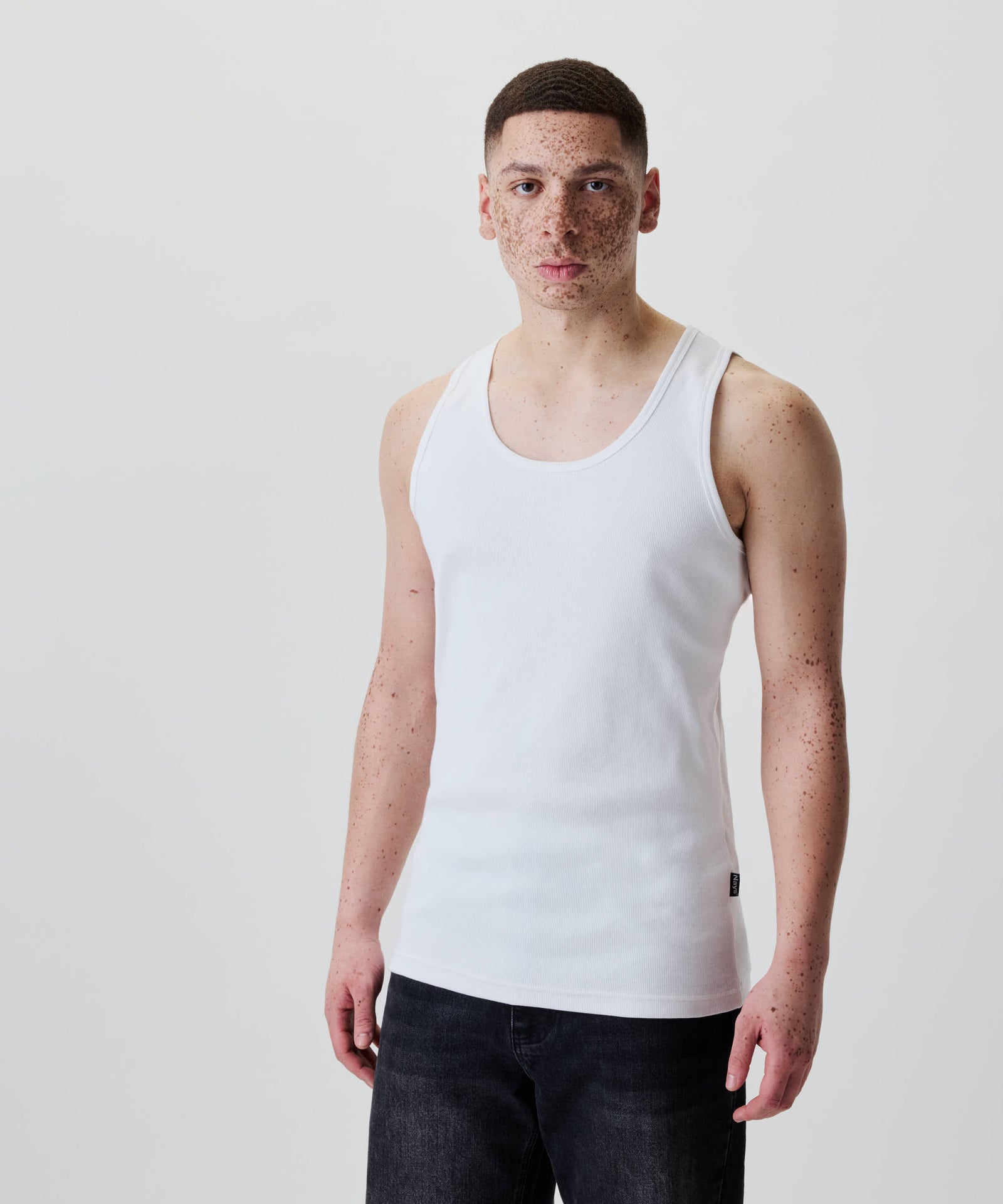 Junger Mann trägt ein Nays Classic Rib Tank Top aus 100 % Baumwolle, zeigt schlichten Stil und Komfort, kombiniert mit dunklen Jeans, neutraler Ausdruck.