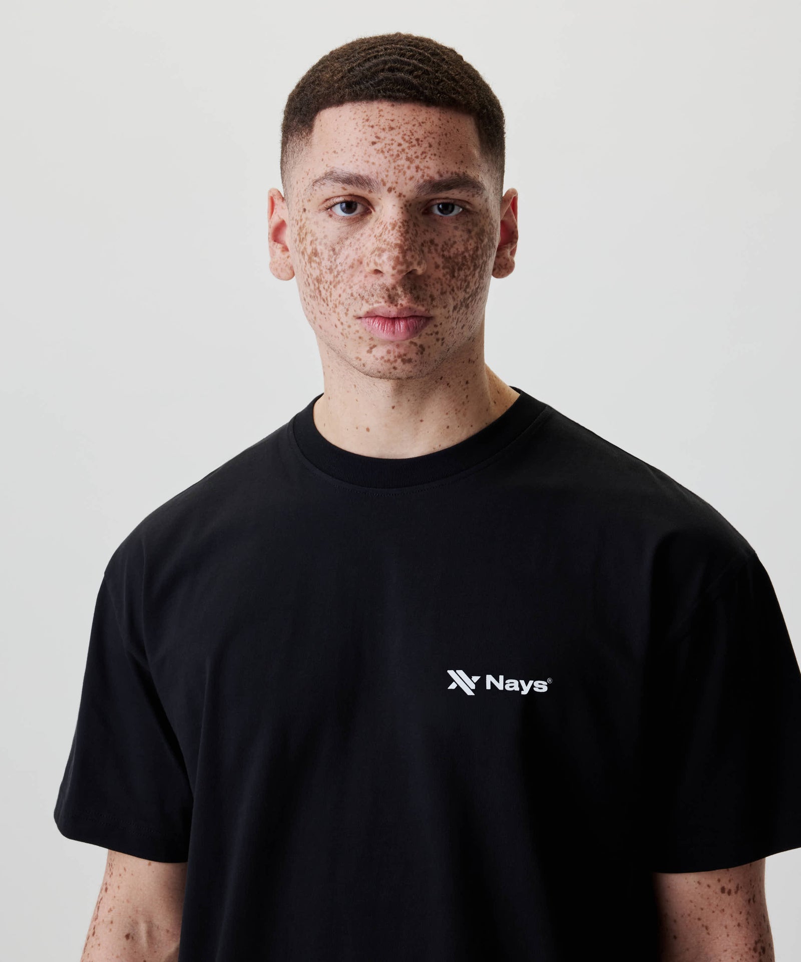 Ein junger Erwachsener trägt das Nays Loose Fit Chest Print Tee mit weißem Logo, entspannter Passform und aus 100% Baumwolle.
