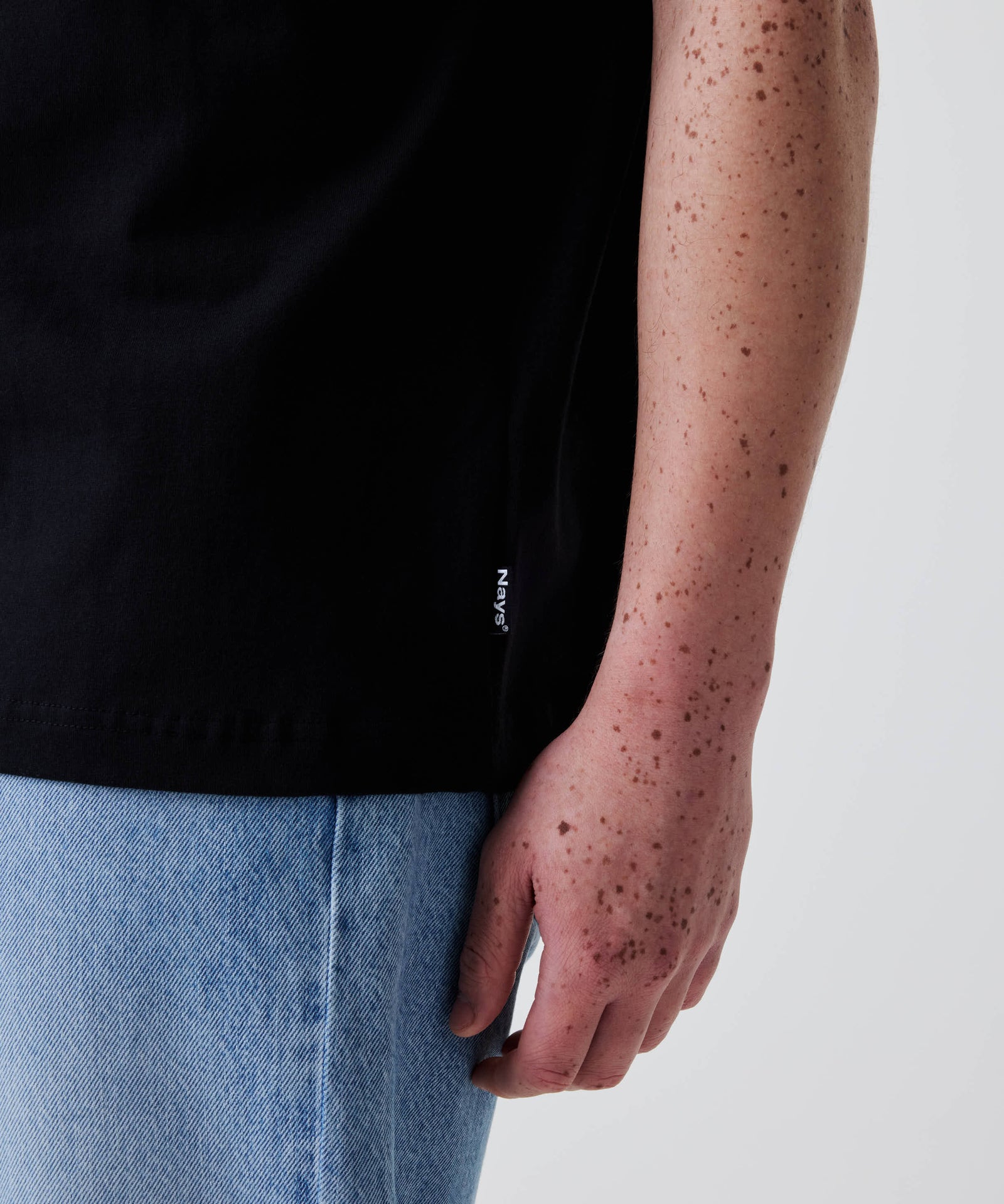 Nahaufnahme eines rechten Unterarms mit Sommersprossen, der das Nays Classic Regular Flag Tee in Schwarz mit kleinem Flag-Label am Saum trägt.