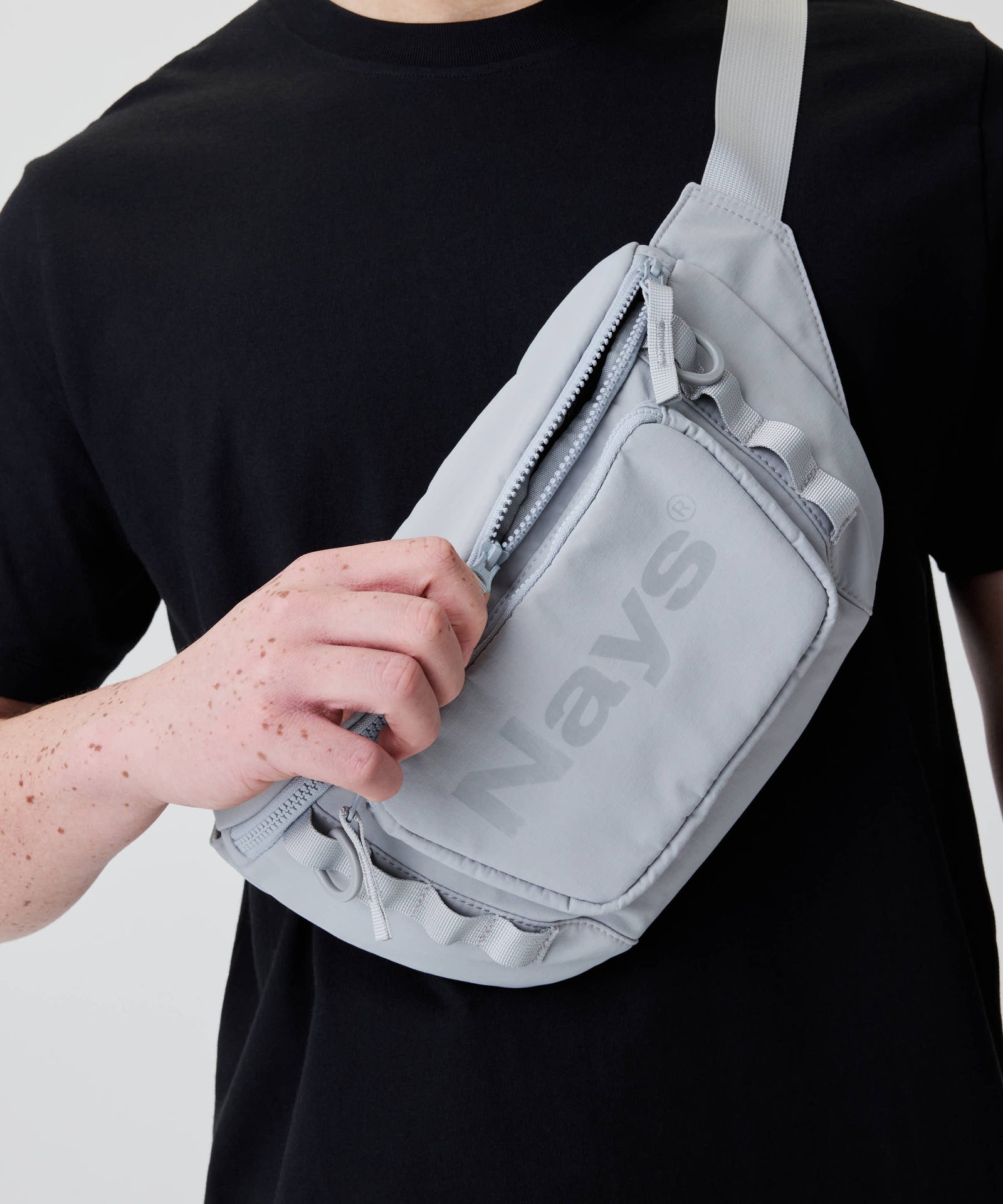 Person mit schwarzem T-Shirt trägt die Nays Lightweight Cross Body Bag aus Nylon, öffnet eine Fronttasche. Tasche mit verstellbarem Gurt und „Nays“ Logo.