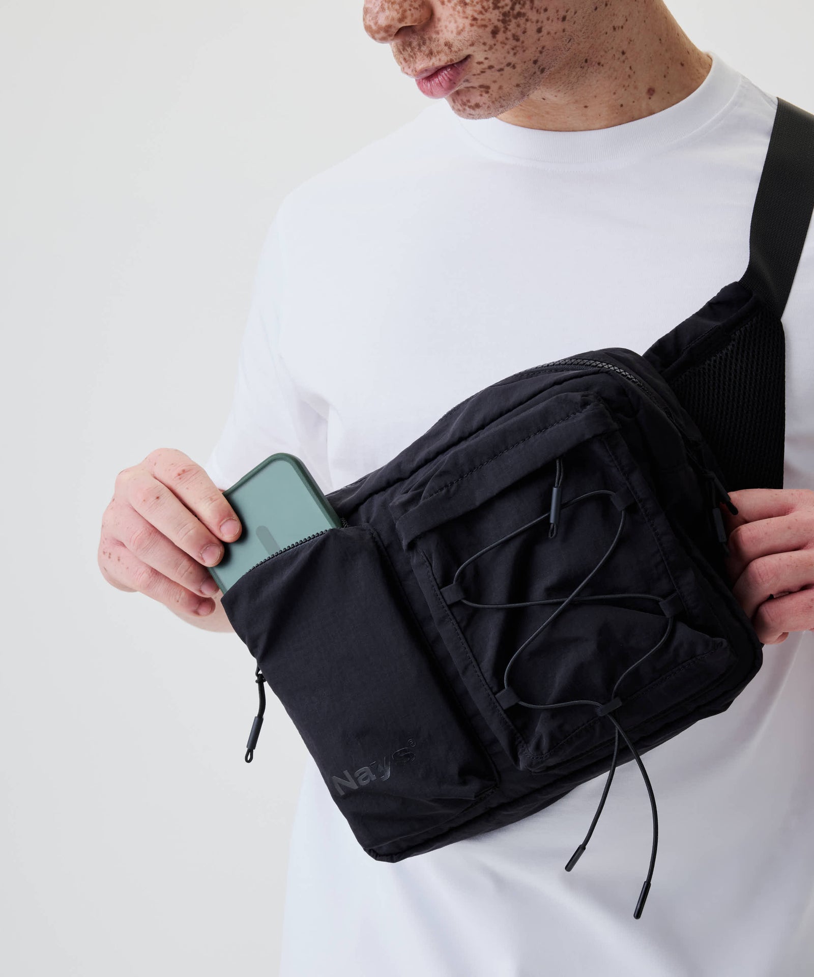 Person mit weißem T-Shirt trägt die Nays Utility Cross Body Bag 2.0, öffnet Fronttasche und steckt ein grünes Smartphone hinein.