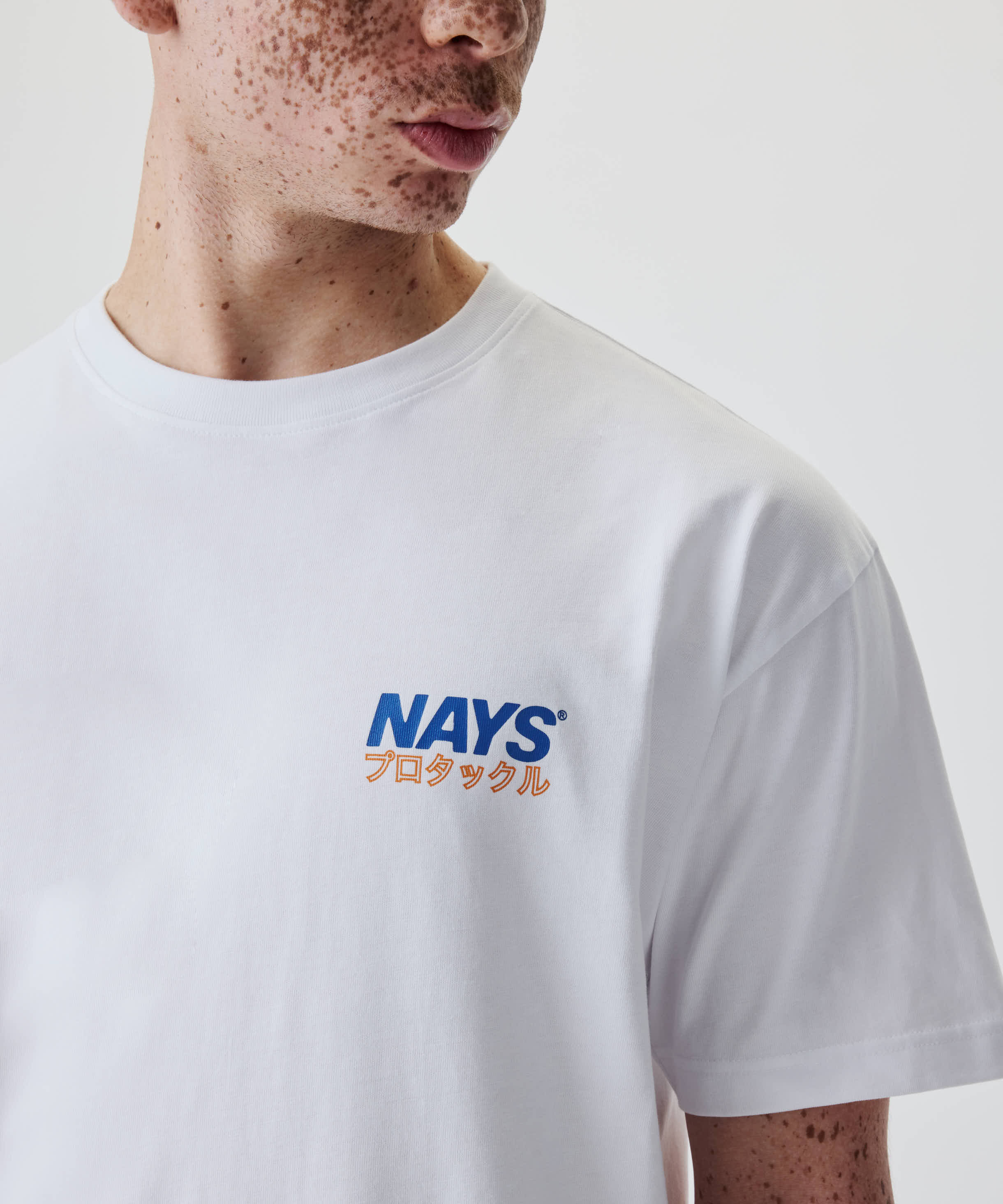 Person trägt das Nays Pro Tackle International Tee, mit freiem Logo und japanischem Schriftzug auf der Brust. Hergestellt aus 100% Baumwolle für Komfort.