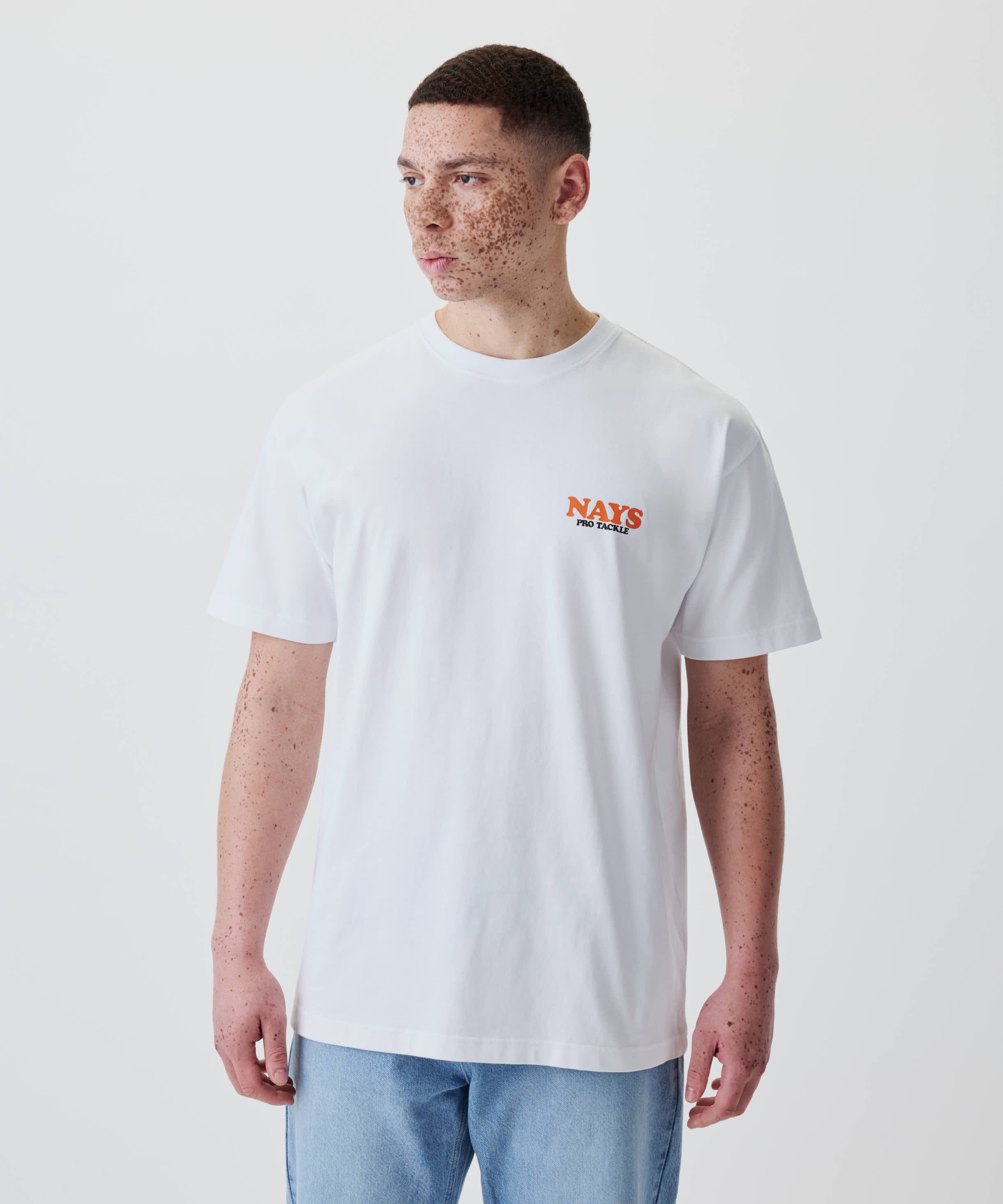 Junger Mann trägt das Nays College Print Tee in Weiß mit kleinem Brustlogo, kombiniert mit hellblauer Jeans. 