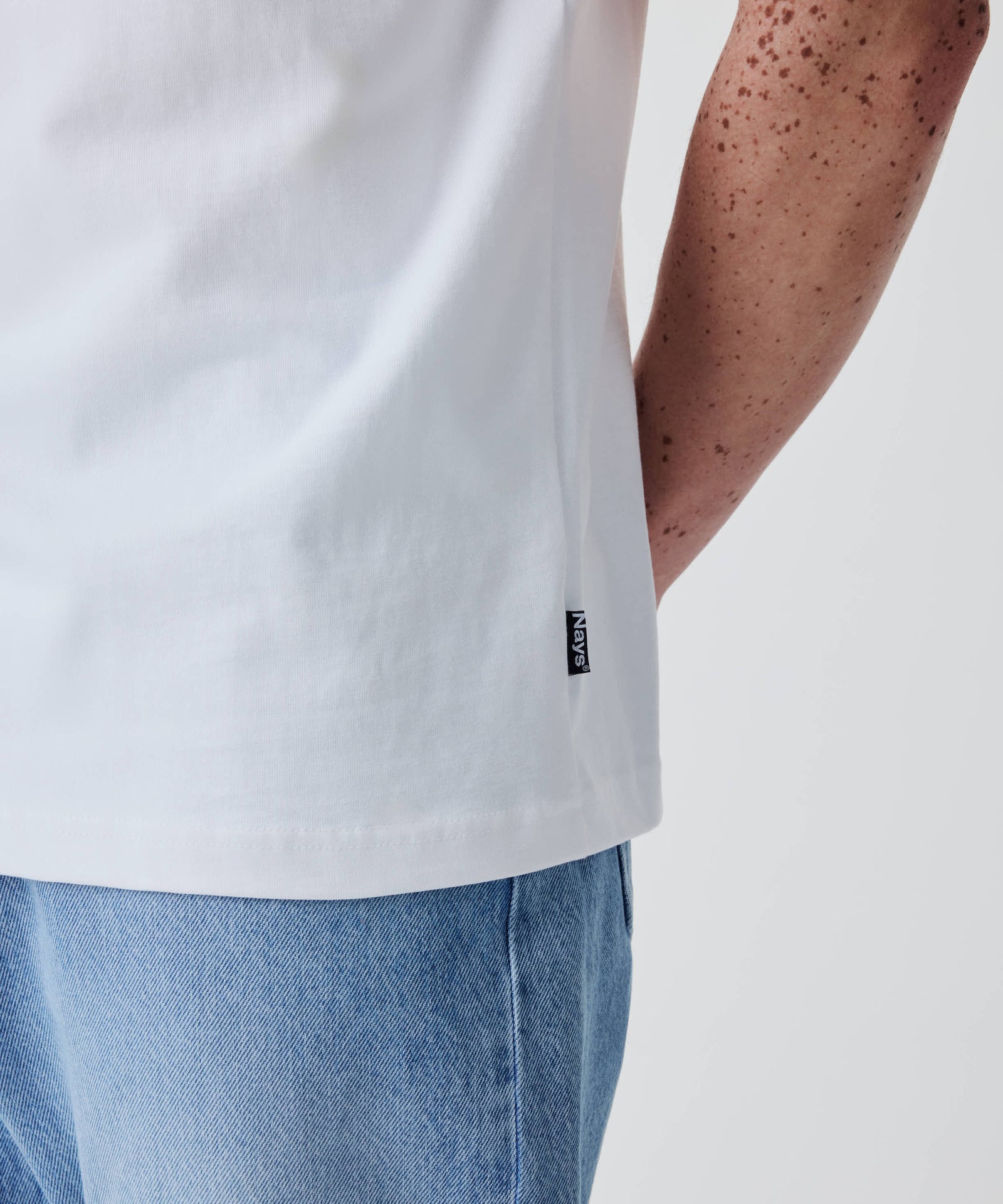 Person im Nays Classic Regular Flag Tee in Weiß, kombiniert mit hellblauen Jeans. Das T-Shirt zeigt einen kleinen schwarzen Stoffanhänger am Saum.